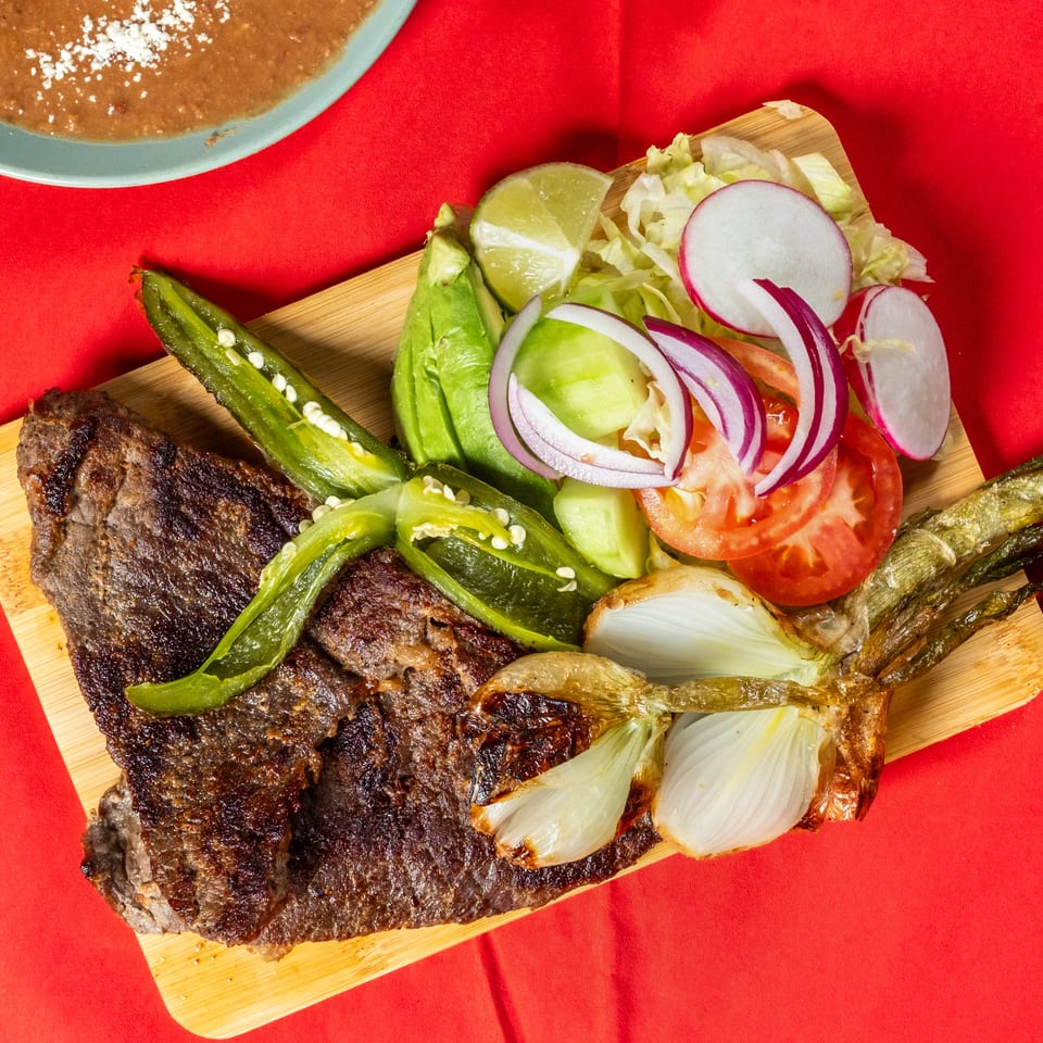 Grilled meat platter with roasted peppers, onions, avocado, lettuce, tomato, radishes, lime, and beans on a wooden board.