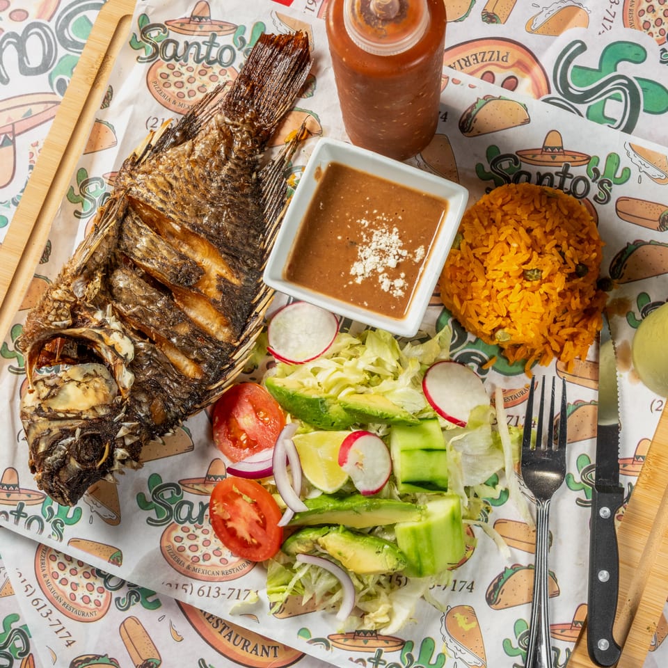 Overhead view of grilled whole fish with orange rice, beans, salad, sauces, and beverages on a colorful Mexican restaurant table.