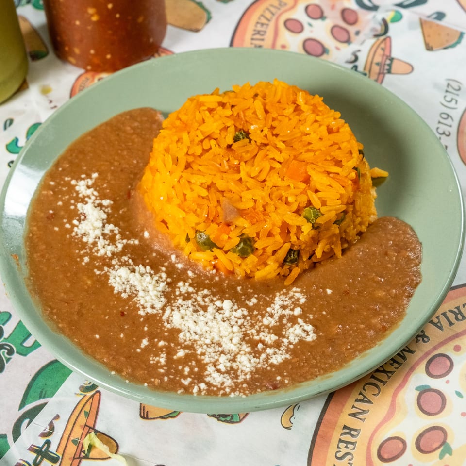 Mexican plate featuring orange vegetable rice, refried beans topped with crumbled cheese, and hot sauce on a colorful tablecloth.