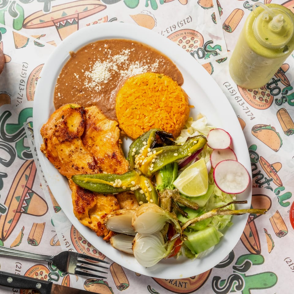 Mexican plate with grilled chicken, orange rice, refried beans, roasted vegetables, salad, radishes, and lime, served with a green drink.