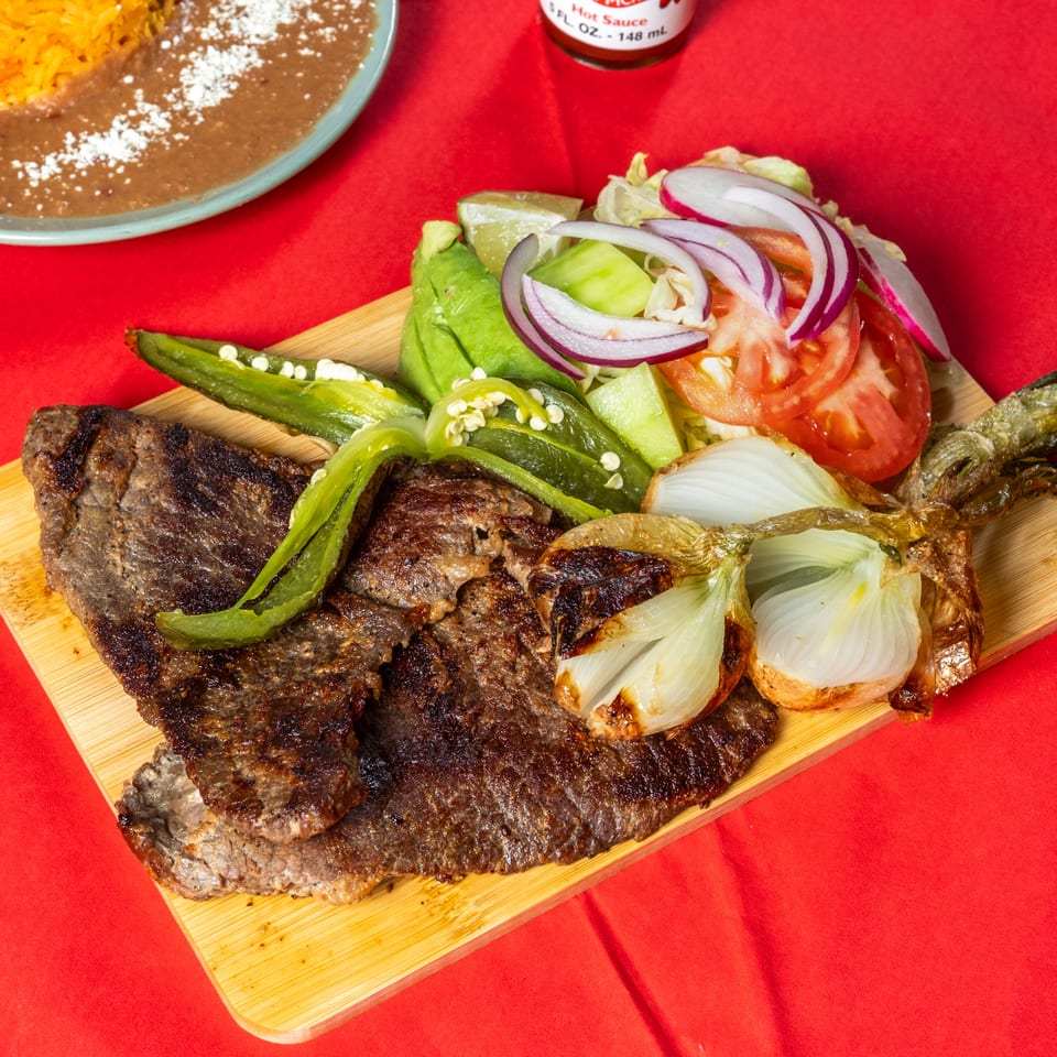 Grilled beef steaks and vegetables served on a wooden platter with salad, rice, beans, lime, and hot sauce.