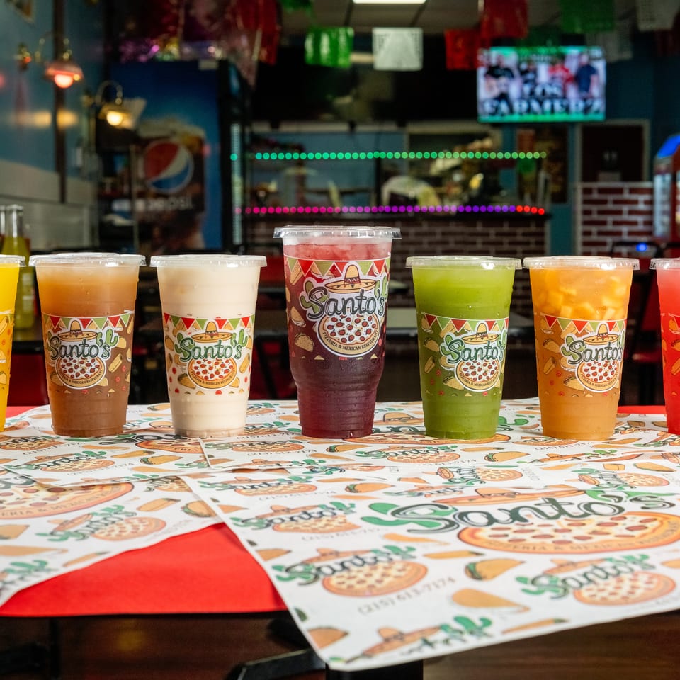 Seven colorful drinks in branded cups line a table inside Santo’s pizzeria and Mexican restaurant.